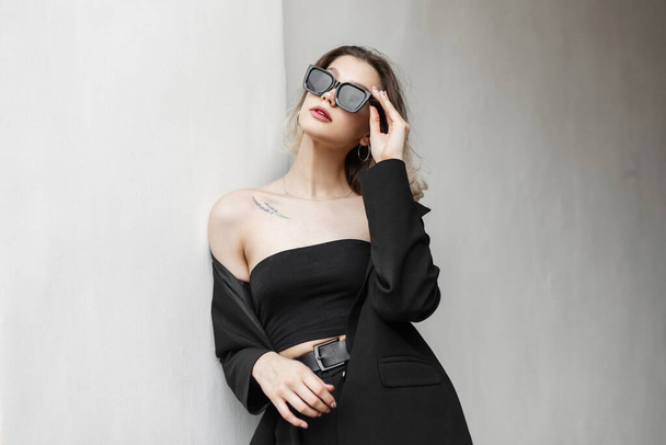 Stylish beautiful elegant vogue woman in a fashionable business urban outfit with blazer and fashion top wearing sunglasses and standing near the white wall on the street - Zdjęcie, obraz