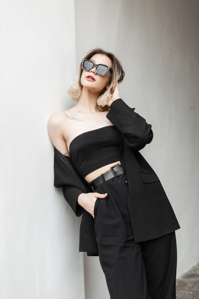 Stylish beautiful elegant girl in fashionable black clothes with trendy top, fashion black blazer and pants standing near the white wall on the street. Women's Urban Business Style - Foto, Imagem