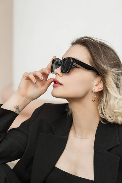 Fashionable young business elegant woman model with trendy modern sunglasses in black outfit with top and blazer sits on the street. Urban female look clothes - Fotografie, Obrázek