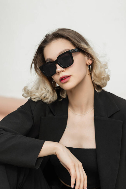 Vogue elegant business fashion beauty woman with fashionable sunglasses in black outfit with top and blazer sits on the street - Φωτογραφία, εικόνα