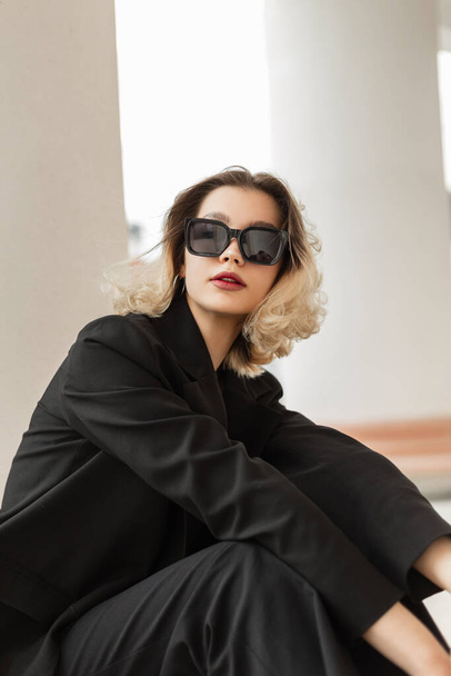 Urban beautiful young business woman model with short hair in black elegance clothes with blazer sits on the street near a white column - 写真・画像