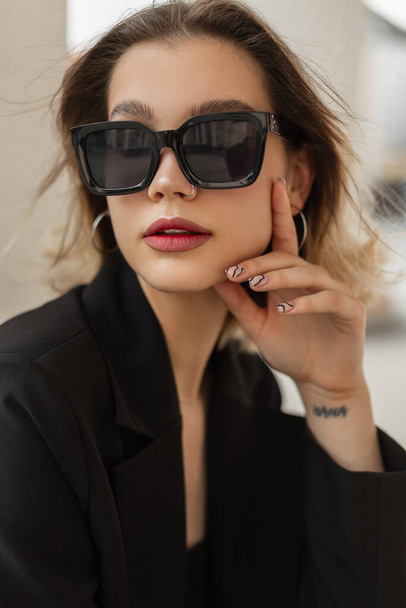 Fresh urban female portrait of business young stylish trendy woman model with cute face with stylish modern sunglasses in black elegant clothes with blazer sits and poses in the city - Fotoğraf, Görsel