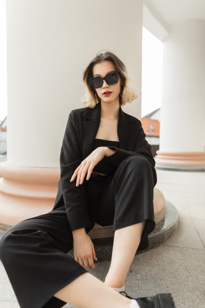 Fashionable young beautiful business woman model with trendy modern sunglasses in black elegant clothes with blazer, top and pants sits near a vintage column in the city - Fotó, kép