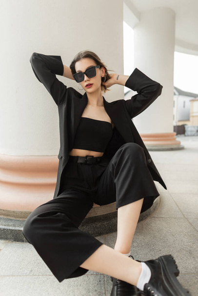 Μοντέρνο μοντέλο νεαρή όμορφη επιχειρηματική γυναίκα με μοντέρνα μοντέρνα γυαλιά ηλίου σε μαύρο κομψό ρούχα με σακάκι, κορυφή και παντελόνι κάθεται κοντά σε μια vintage στήλη στην πόλη - Φωτογραφία, εικόνα