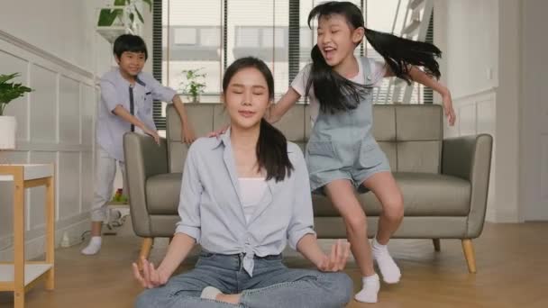Young Asian Thai mother sits on the living room floor, meditates, and practices yoga for health and wellness, the children play, chaotic and naughty, happy domestic home lifestyle on family weekend. - Felvétel, videó