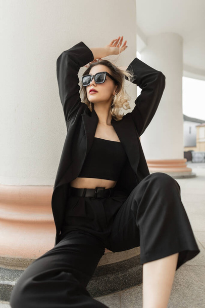 Μόδα αξιολάτρευτη όμορφη νεαρή κομψότητα γυναικείο μοντέλο με κομψά γυαλιά ηλίου σε μαύρο επιχειρηματικό μοντέρνα ρούχα με σακάκι και κορυφή κάθεται και ποζάρει στην πόλη κοντά σε μια vintage λευκή στήλη - Φωτογραφία, εικόνα