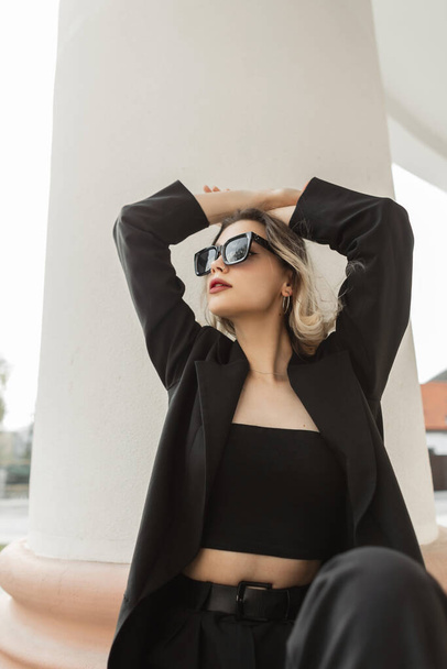 Stylish beautiful elegant girl with cool sunglasses in fashionable black business clothes with blazer and top sitting and posing near a white vintage column in the city - Foto, Bild