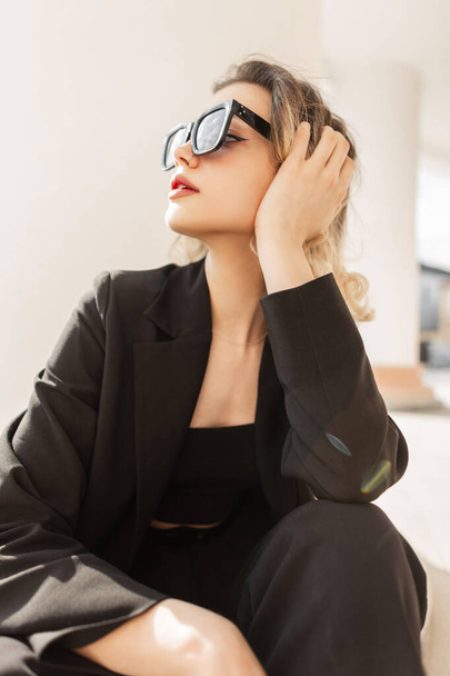 Fashion glamour pretty lade woman with modern sunglasses in black elegant business clothes sits and rest on the street - Fotoğraf, Görsel
