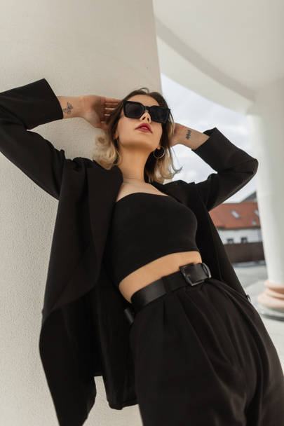 Fashionable beautiful elegant business girl in fashionable black clothes posing near a white vintage column - Zdjęcie, obraz
