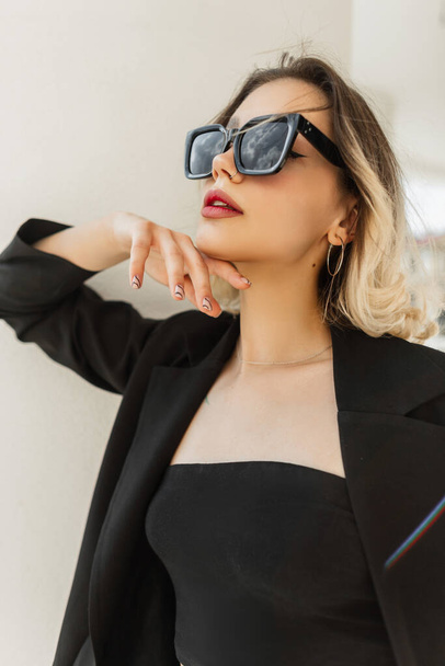 Красивая молодая элегантная стильная женщина модель хипстера в модных черных солнцезащитных очках с черным бизнес-блейзер и стоя у белой стены на улице - Фото, изображение