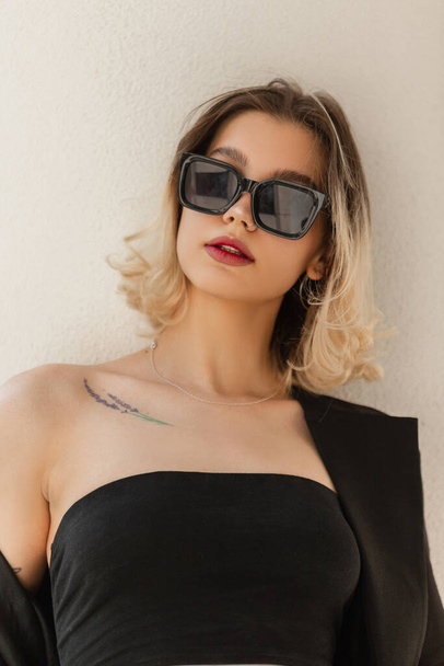 Woman's fresh urban portrait of a beautiful young woman with short hair with cool black sunglasses and a black top with blazer stands near a white wall in the city - Fotografie, Obrázek