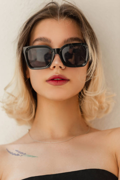 Urban female summer fresh portrait of beautiful young woman model with cool sunglasses in black top stand on a white background on the street - Photo, Image