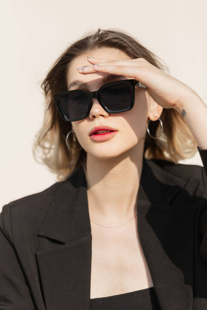 Fashionable beautiful business lady model with modern sunglasses in fashion black elegant clothes with stylish blazer sits in the sunny day and covers her eyes with hand. Summer female urban look - Foto, imagen