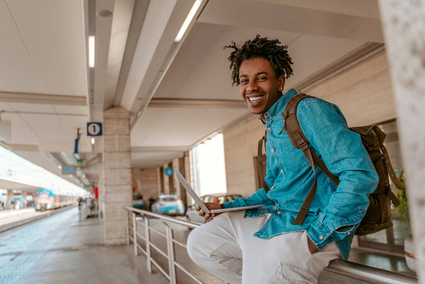 Good day. Smiling african american guy with backpack on back sitting with laptop near railroad looking at camera - Fotoğraf, Görsel