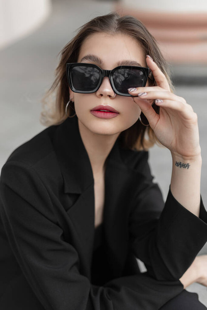 Elegance fashion stylish business beauty woman in black fashionable clothes with blazer wears a trendy modern sunglasses and sits on the street - Fotó, kép