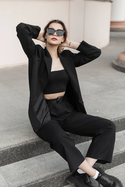 Trendy fashionable young beautiful woman model with modern sunglasses in elegant business urban outfit with top, blazer, pants and shoes sits on the steps on the street - Zdjęcie, obraz