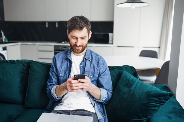 man sitting at home, sending messages on smartphone, smiling, sitting on green sofa in bright living room - Photo, Image