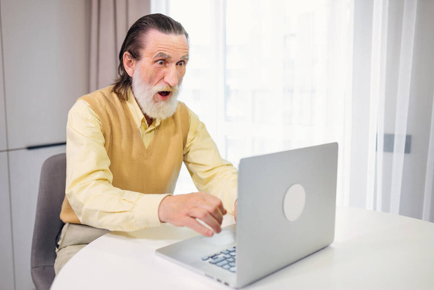 ショックを受けた、ストレスを受けた高齢者はノートパソコンを見て、自宅で信じられないほどのオンラインニュースを読み取り、老人の祖父は予期せぬ問題に驚いています - 写真・画像