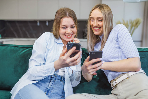 Χαρούμενες φίλες που τσεκάρουν φωτογραφίες σε ένα κινητό τηλέφωνο, που περνούν χρόνο μαζί στο σπίτι, Δύο νεαρές κυρίες κάθονται δίπλα-δίπλα σε έναν καναπέ στο σαλόνι, χρησιμοποιώντας ένα smartphone - Φωτογραφία, εικόνα