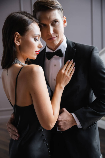 Brunette woman in dress touching boyfriend in formal wear at home  - 写真・画像