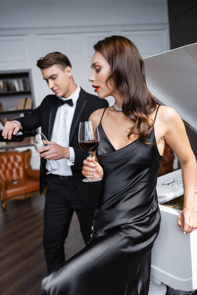Sensual woman in dress holding glass of wine near blurred boyfriend and piano at home  - Foto, Imagen