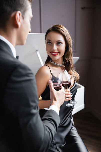 Χαμογελαστή γυναίκα με μεταξωτό φόρεμα που κρατά ένα ποτήρι κρασί κοντά στον θολή φίλο της με κοστούμι στο σπίτι  - Φωτογραφία, εικόνα