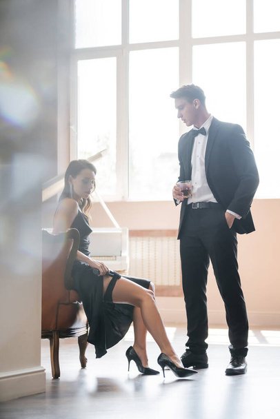 Elegant man holding glass of whiskey near sensual girlfriend putting gun in garter at home  - Valokuva, kuva