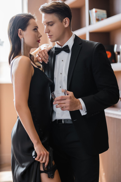 Sensual woman holding handgun and looking at boyfriend in suit holding glass of whiskey  - Fotó, kép