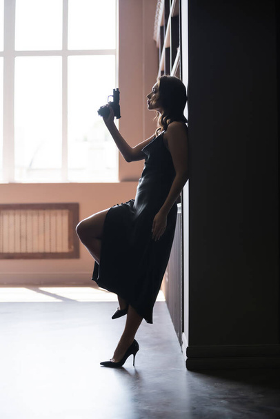 Side view of sensual woman in dress holding handgun at home  - Foto, afbeelding