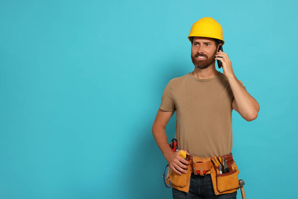 Professional builder in hard hat with tool belt talking on phone against light blue background, space for text - Fotó, kép