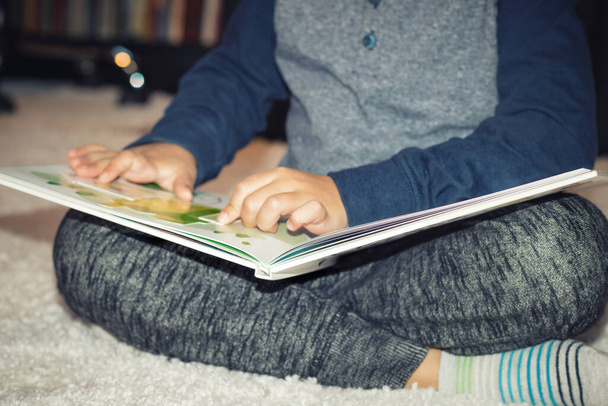 Маленький мальчик читает историю на ковре. Мальчик читает книгу, сидя на полу.  - Фото, изображение