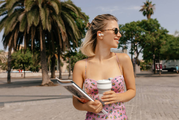 Κομψό νεαρή καυκάσια γυναίκα σε γυαλιά ηλίου με καφέ και περιοδικά που ποζάρουν στο δρόμο στέκεται σε εξωτικά φυτά φόντο. Επιτυχημένη αξιολάτρευτη κυρία περνούν χρόνο σε εξωτερικούς χώρους - Φωτογραφία, εικόνα