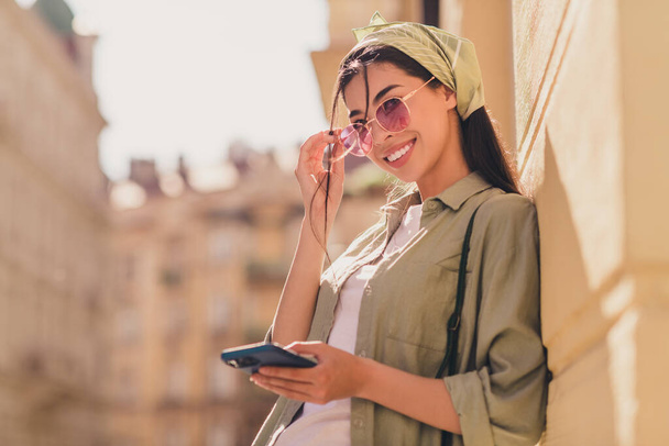 Portrait of attractive cheerful lady toothy smile hand touch sunglass hold telephone walk city center outside. - 写真・画像