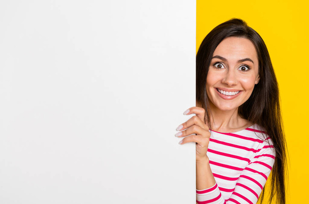 Photo portrait of charming young woman stand behind blank white empty space dressed trendy striped look isolated on yellow color background. - 写真・画像
