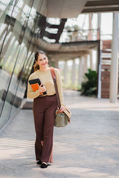 Азиатка, держащая планшет и сумку, идет рядом со зданием образовательной концепции - Фото, изображение