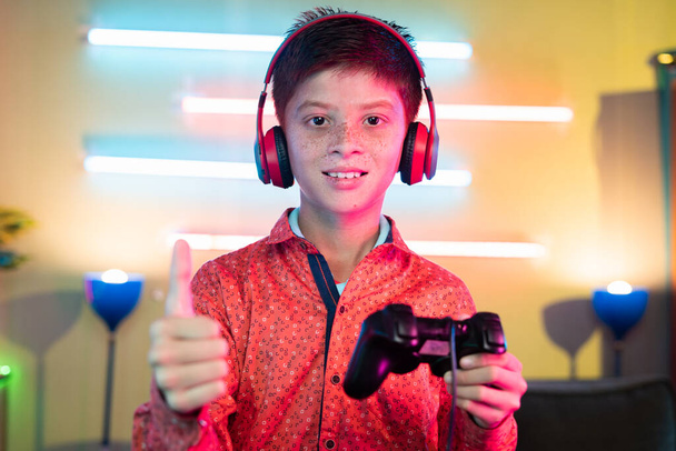 young Teenager kid with headphones holding gamepad by showing thumbs up by looking at camera at home - concept of live streamer and gaming vlogger - Photo, Image