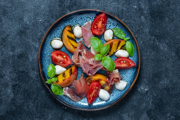 Grilled peach salad with mozzarella, prosciutto ham, red tomato, green basil and soy sauce. Fresh gourmet salad in plate on black background, close up, top view, copy space - Photo, Image