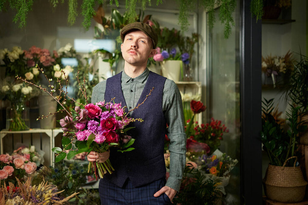 Serious emotion caucasian male in peaked cap and vintage clothes from 20s holding beautiful bouquet of red and violet flowers. Congratulating with flowers. Mothers Day or Valentines Day concept - Zdjęcie, obraz
