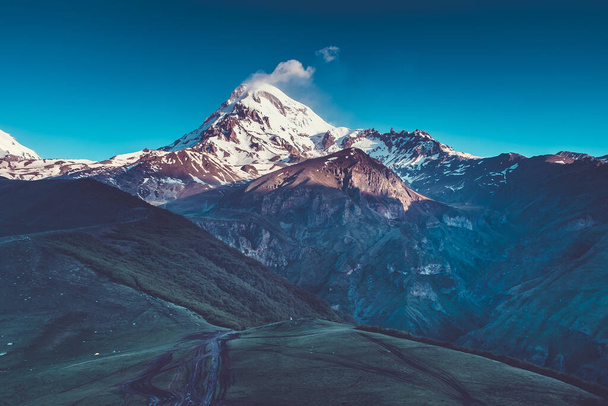 Panoramic landscape of snow-capped mountain Kazbek at dawn and tourist trails on green valley below. Beautiful sunrise. Blue toning - Valokuva, kuva