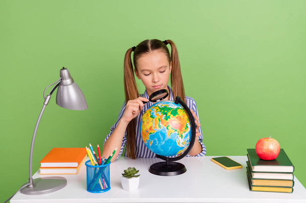 Photo of serious interested small schoolkid dressed striped uniform tails enlarging globe sitting desktop isolated green color background. - Foto, Imagen