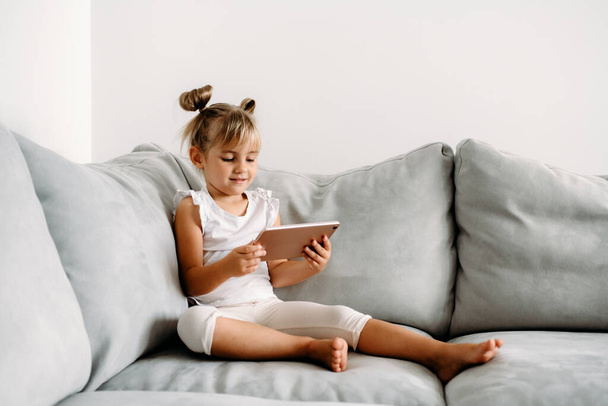 Κοριτσάκι που παίζει με ψηφιακό ασύρματο tablet υπολογιστή στον καναπέ στο σπίτι. Βρεφική ανάπτυξη με online εφαρμογές. Η έννοια των παιδικών και ηλεκτρονικών συσκευών. Πορτρέτο του παιδιού με smartphone. Εκπαιδευτική και μαθησιακή έννοια. - Φωτογραφία, εικόνα