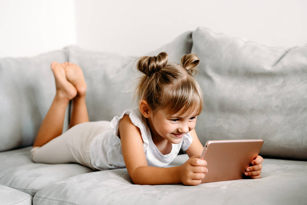 Κοριτσάκι που παίζει με ψηφιακό ασύρματο tablet υπολογιστή στον καναπέ στο σπίτι. Βρεφική ανάπτυξη με online εφαρμογές. Η έννοια των παιδικών και ηλεκτρονικών συσκευών. Πορτρέτο του παιδιού με smartphone. Εκπαιδευτική και μαθησιακή έννοια. - Φωτογραφία, εικόνα