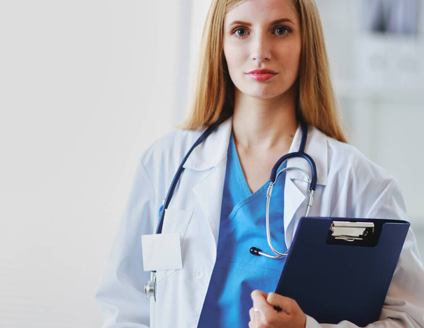 Portrait of woman doctor with stethoscope at hospital corridor, holding a folder - Foto, imagen