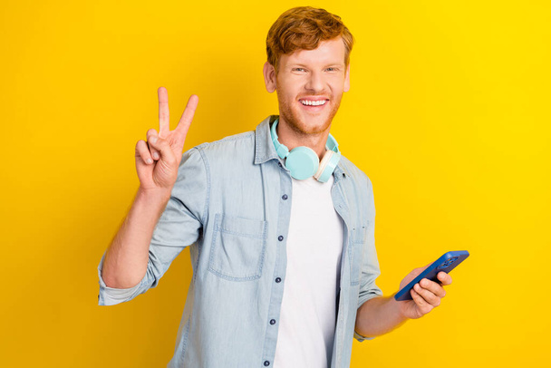 Photo of cheerful positive boy man trendy jeans outfit show v symbol arm hold telephone enjoy playlist isolated on yellow color background. - Fotó, kép