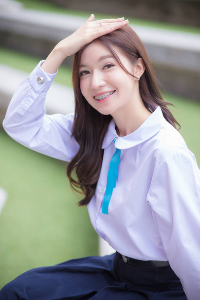 Beautiful Asian high school student girl in the school uniform with braces on her teeth sitting and smiles confidently while she looks at the camera happily with the building in the background. - 写真・画像