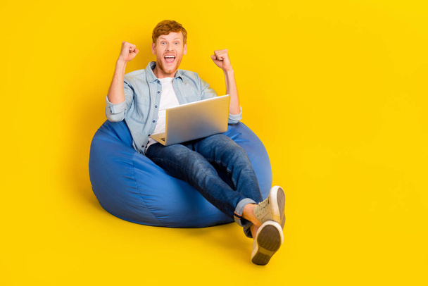 Photo of lucky funny guy dressed denim shirt sitting bean bag reading modern device fists empty space isolated yellow color background. - Фото, зображення