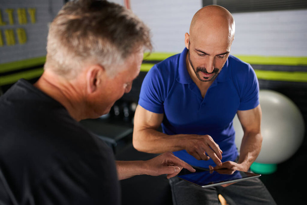 Male physio therapist and wellness personal trainer having a consultation with elderly senior man on strength and rehabilitation and assisted exercise routine in home gym looking at digital tablet - Foto, imagen