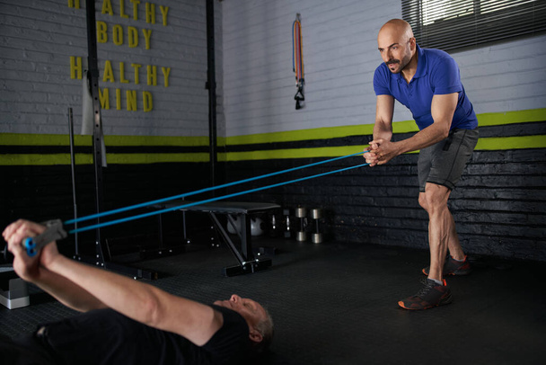 Male personal trainer holds onto elastic rubber resistance bands while an elderly mature man lying on exercise mat on floor and does bicep curl for strength and rehabilitation exercise - Foto, immagini