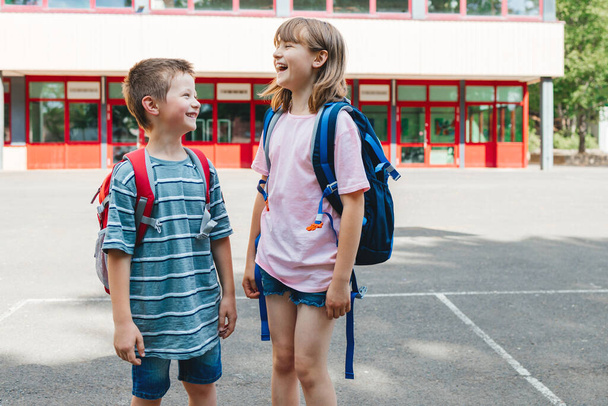 Хлопчик і дівчинка, брат і сестра стоять з рюкзаками на спині перед будівлею школи і сміються. Діти повертаються до школи після канікул
. - Фото, зображення