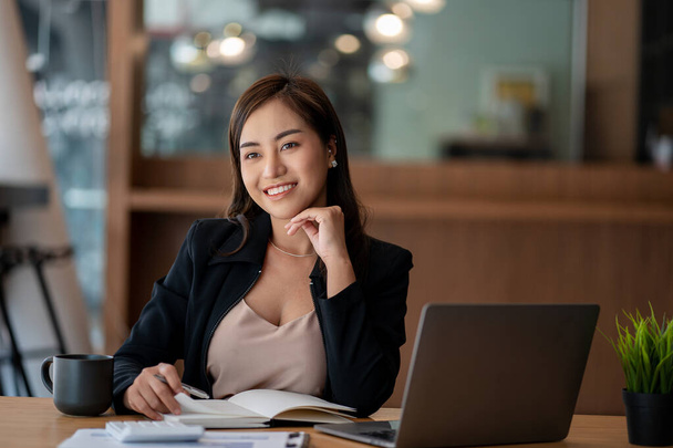 Азиатская деловая женщина или бухгалтер, работающая с ноутбуком и мобильным телефоном. Улыбайтесь и радуйтесь, сидя за своим столом с финансовыми документами. современный офисный калькулятор - Фото, изображение